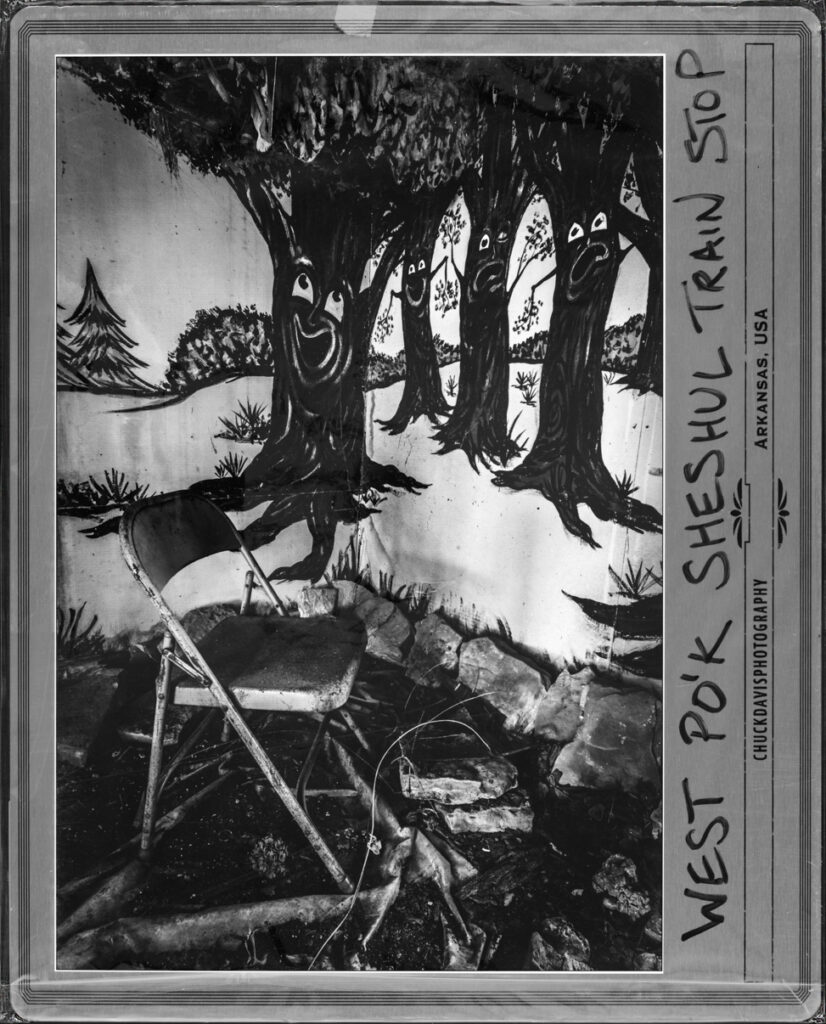 Black-and-white fine art photograph of an abandoned train stop scene in Arkansas featuring a weathered chair, stone landscaping, and painted tree mural.